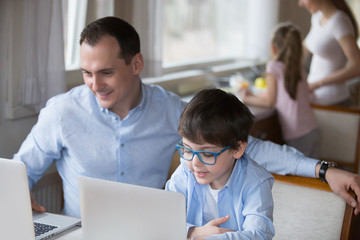 Happy family with small children spend free time together at home. Little son copying father sitting at the table using computers reading news having fun, mother and daughter in kitchen on background