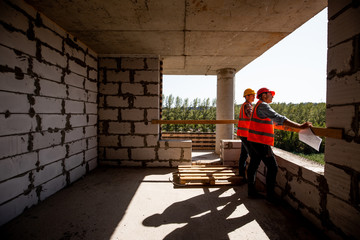 Two young civil engineers dressed in orange work vests and helmet stand on the building site inside the building under construction