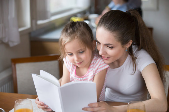 Attractive woman holds book sitting together with adorable little girl indoors. Young loving mother read fairy tale to pretty preschool daughter. Happy family spend free time together at home concept