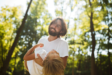 Dark-haired father with beard dressed in the white t-shirt is holding in the arms his blond son and smiling on a summer day in the park.