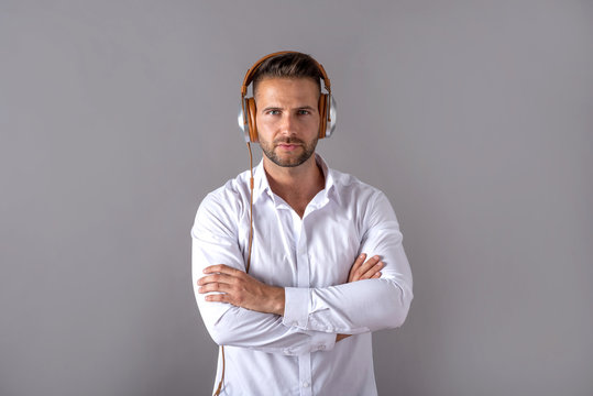 A serious handsome young man in a white shirt listening to music on his headphones and standing in front of a grey background in the studio.