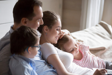 Obraz premium Mother father little daughter and son relaxing sitting on sofa at home spending free time on weekend together watch movie cartoon funny videos have fun surfing internet. Happy friendly family concept