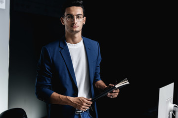 Stylish young architect in glasses dressed in a blue checkered jacket keeps a notebook in his hands in the office