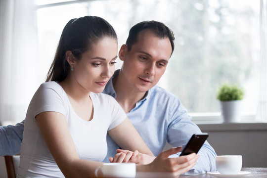 Millennial positive wife and husband sitting together in kitchen spend free time at home hold mobile phone reading email good news and messages. Spouses making order via internet online using gadget
