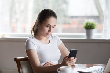 Fototapeta premium Pensive millennial female sitting in kitchen at table hold smartphone read email, browsing surfing internet. Serious woman use mobile phone feels frustrated sad upset receive unpleasant message news