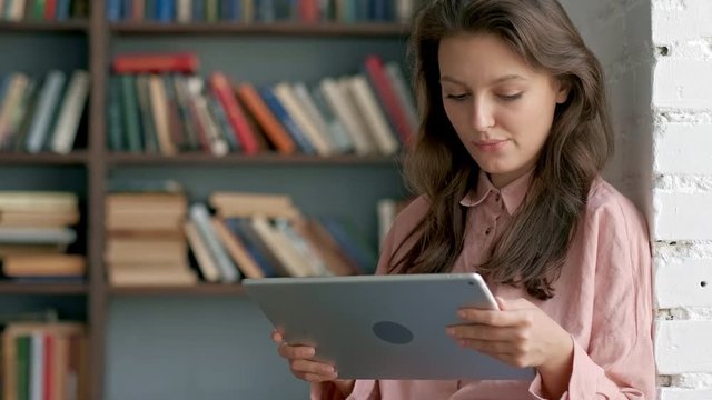 Close up of the curly young woman in glasses working on the tablet device and smiling to the camera at the library passage. Portrait. Indoors