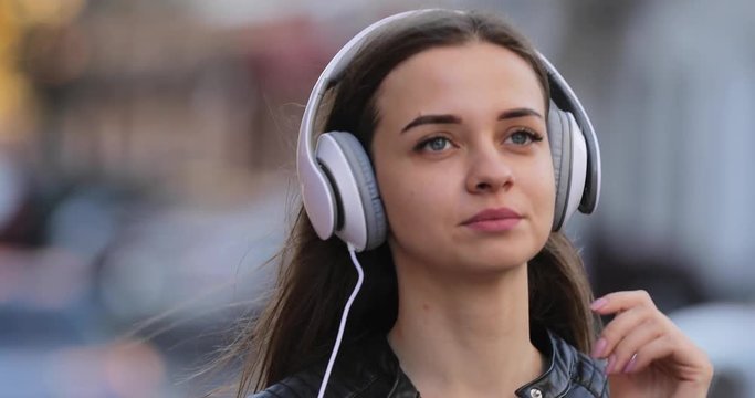 Modern woman puts on headphones while walking along city street, listening music