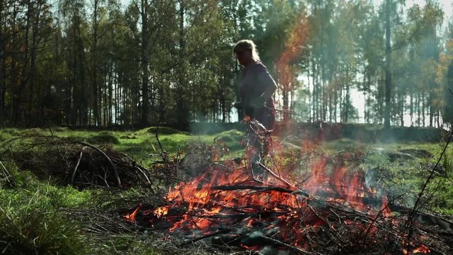 Bonfire in autumn. Bonfire on a meadow to get rid of old bushwood. Woman throwing old branches and leaves on fire.