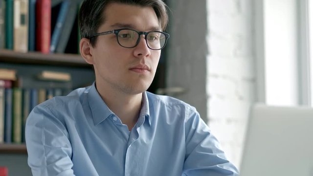 College student works on essay at the library. Attractive Asian man with glasses reading book and typing on computer. 4k