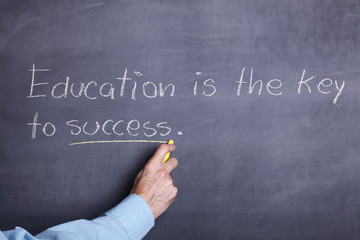 A man's hand holding a piece of chalk and writing the phrase: "Education is the key to success" on a blackboard