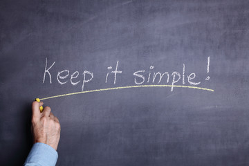 A man's hand holding a piece of chalk and writing the phrase: "Keep it simple!" on a blackboard