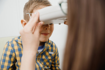 Little blonde boy in plaid shirt looks at ophthalmologist through special device to check vision modern clinic
