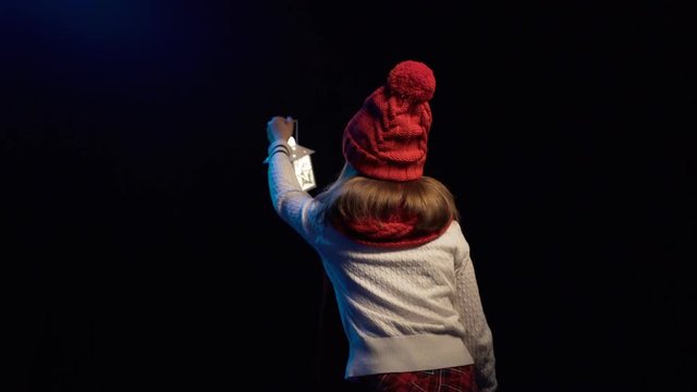 Back view of a little girl wearing knitted winter hat and scarf standing with lantern searching looking to sides, over black background