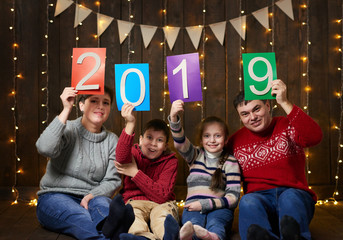 family posing with 2019 new year text, sitting on dark wooden background with christmas lights and...