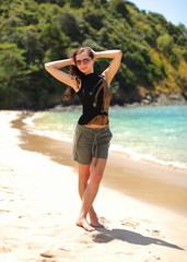 Fototapeta premium Young woman in black t shirt, shorts and sunglasses, standing on the beach, hands in her hair, sea and tree covered hills in background.
