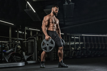 Young handsome male athlete bodybuilder weightlifter with idial abdominals, doing exercises in modern gym on a dark background. Exercises for biceps.  - 230498348