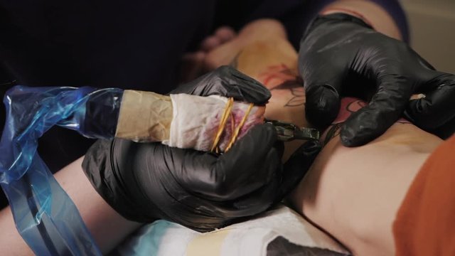 Professional master is painting tattoo with red ink, fills drawing with color. Works in black latex gloves with handmade rotor gun machine in studio. Slow motion