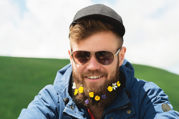 Close-up portrait of a cheerful bearded man in sunglasses and a gray cap with wildflowers in a...