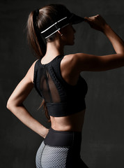 Young sport woman in workout exercise cloth and cap posing stretching in gym on dark grey background