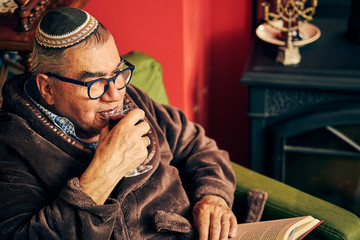 Jewish senior with glasses sitting in the armchair reading a torah book and drinking kosher wine