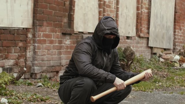 Bandit man in black mask and jacket with hood with baseball bat sits on street