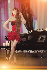 Beauty young model woman in luxurious evening red dress in a fairy luxury apartment. Beautiful makeup and lond hair. Classic vintage home interior. Light rays of a nightclub.