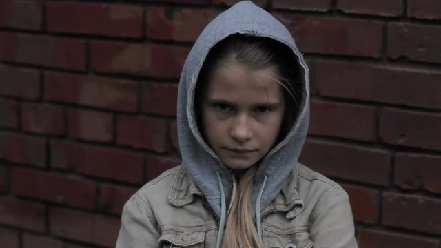 Dark portrait of sad displeased young girl looking at camera against brick wall. Difficult child wearing hooded jacket feeling of anger for having been offended standing alone outdoor.