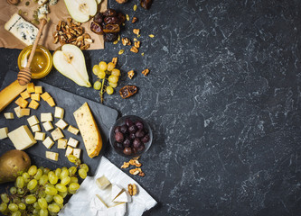 Different types of cheese on board, olive, fruits, almond and wine glasses on black stone table