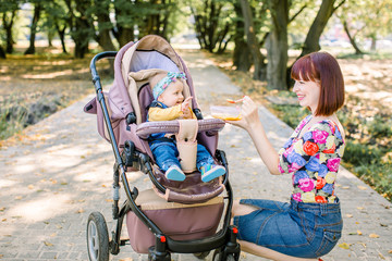 Obraz premium Young attractive mother feeding her cute baby girl, giving her first solid food, healthy vegetable pure from carrot with a plastic spoon sitting on baby stroller carriage and posing smiling