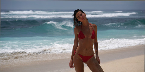 Young woman in red swimwear posing against big waves of the ocean on white sandy beach