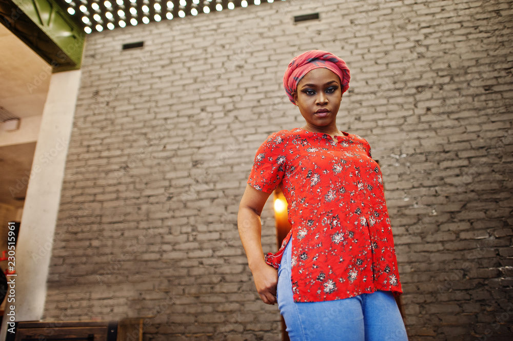 Wall mural Stylish african woman in red shirt and hat posed against brick wall.