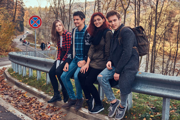 Obraz na płótnie Canvas Group of young friends with backpacks sitting on guardrail near road with a beautiful forest and river in the background.