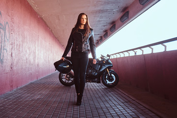 A beautiful biker girl holding helmet next superbike walks on a sidewalk inside the bridge.