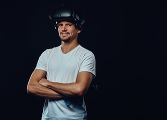 Pro gamer with bristle dressed in white shirt wearing virtual reality glasses and holds joysticks, posing with crossed arms.