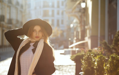 Fototapeta premium Outdoor portrait of awesome blonde girl wearing hat, scarf and coat, posing at the old street in sun glare. Empty space