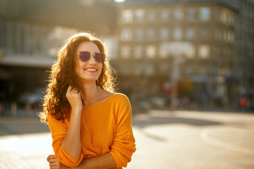 Cheerful young model with curly hair in sunglasses wearing trendy outfit and walking at the sunny street. Space for text