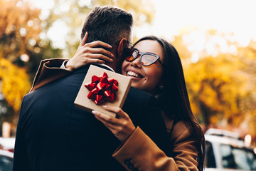 Couple. Love. Holiday. Outdoors. Woman is holding a gift box and smiling while hugging her man in the street