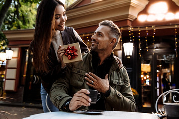 Couple. Holiday. Cafe. Woman is giving a gift box to her man. Both are in warm casual clothes smiling while sitting in the cafe outdoors