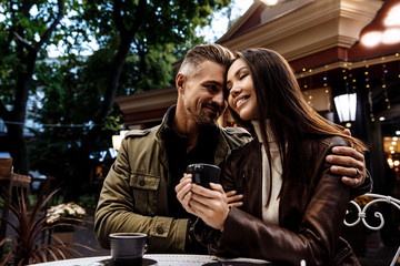 Cafe. Couple. Love. Autumn walk. Man and woman in warm casual clothes are drinking coffee, hugging and smiling while sitting in the cafe outdoors