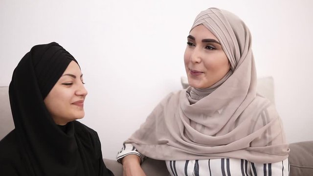 Two pretty muslim woman sitting on sofa relaxing and talking. Women in beige and black headscarf have friendly coversation at home