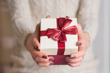 Girl in sweater holding gift box with red ribbon tie bow. Present for Christmas and New Year holiday. Close up, selective focus