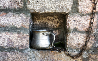 Metal cup for drinking water on a chain in the brick wall