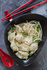 Noodle soup with wontons served in a black bowl, view from above on a grey concrete surface, close-up