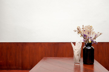 Brown glass bottle with dried white and purple flower and napkin in a glass on wooden table in a retro vintage cafe with white and wooden panel background
