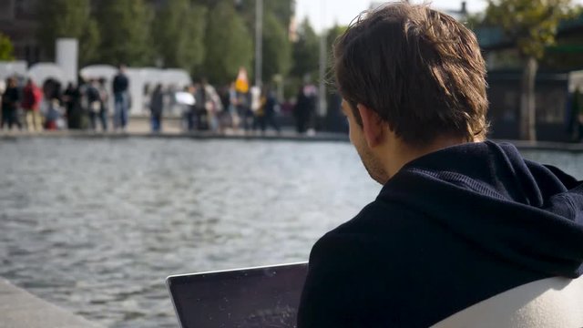 Handsome young man sitting on the bank of a canal in Amsterdam typing on the laptop. Netherlands. Flat plane