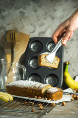 A slice of banana bread in the tongs in the female hand. Homemade cakes among kitchen utensils, bananas and hazelnuts