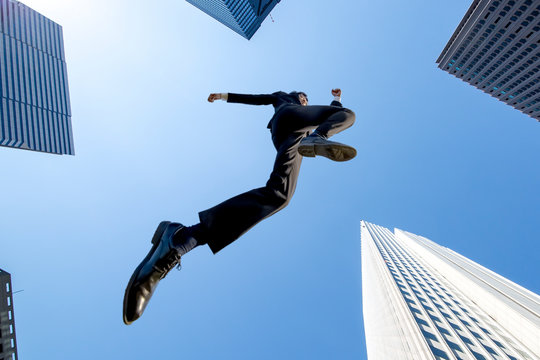 ビル街を青空バックにジャンプするスーツ姿のビジネスマン1人。挑戦,努力,成功,元気イメージ