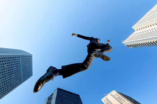 ビル街を青空バックにジャンプするスーツ姿のビジネスマン1人。挑戦,努力,成功,元気イメージ