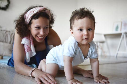Picture of happy emotional young Hispanic female with curly hair spending great time with her toddler son, teaching him how to crawl, smiling, rejoicing at his growth, development and achievements