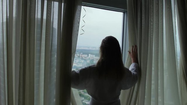Woman in bathrobe unveil curtains in hotel room and look on cityscape through window